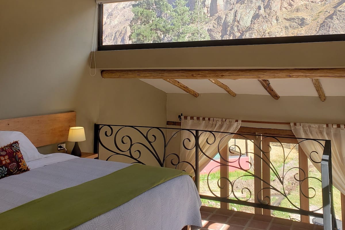 A bedroom with a bed, a nightstand, a lamp, a window with a view of a mountainous landscape, and a balcony with a railing.