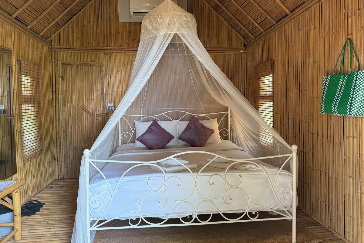 A bed with a white metal frame and a canopy, set in a room with bamboo walls and a sloped ceiling.