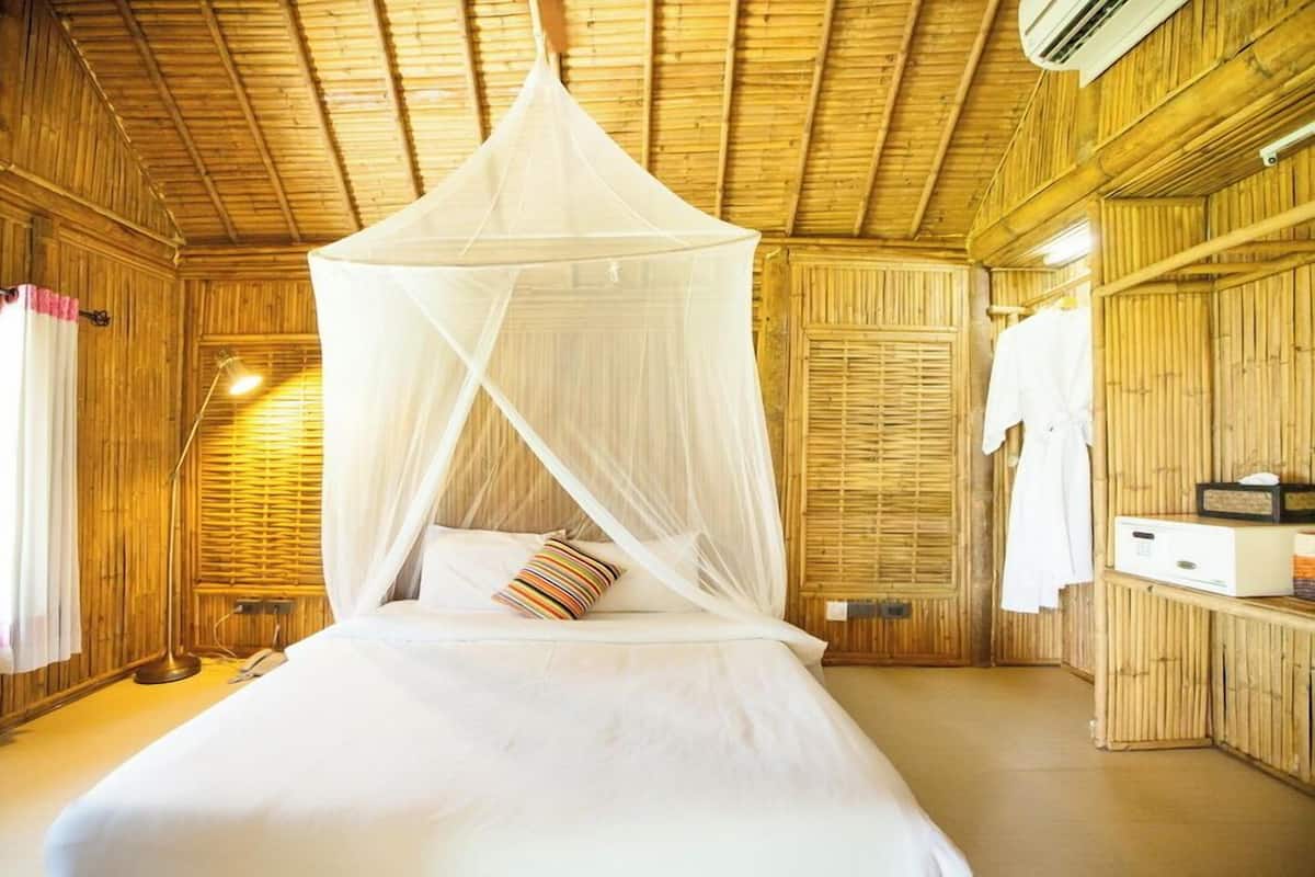 A room with a bed under a white canopy, wooden walls, a bedside table with a radio, and a window with curtains.