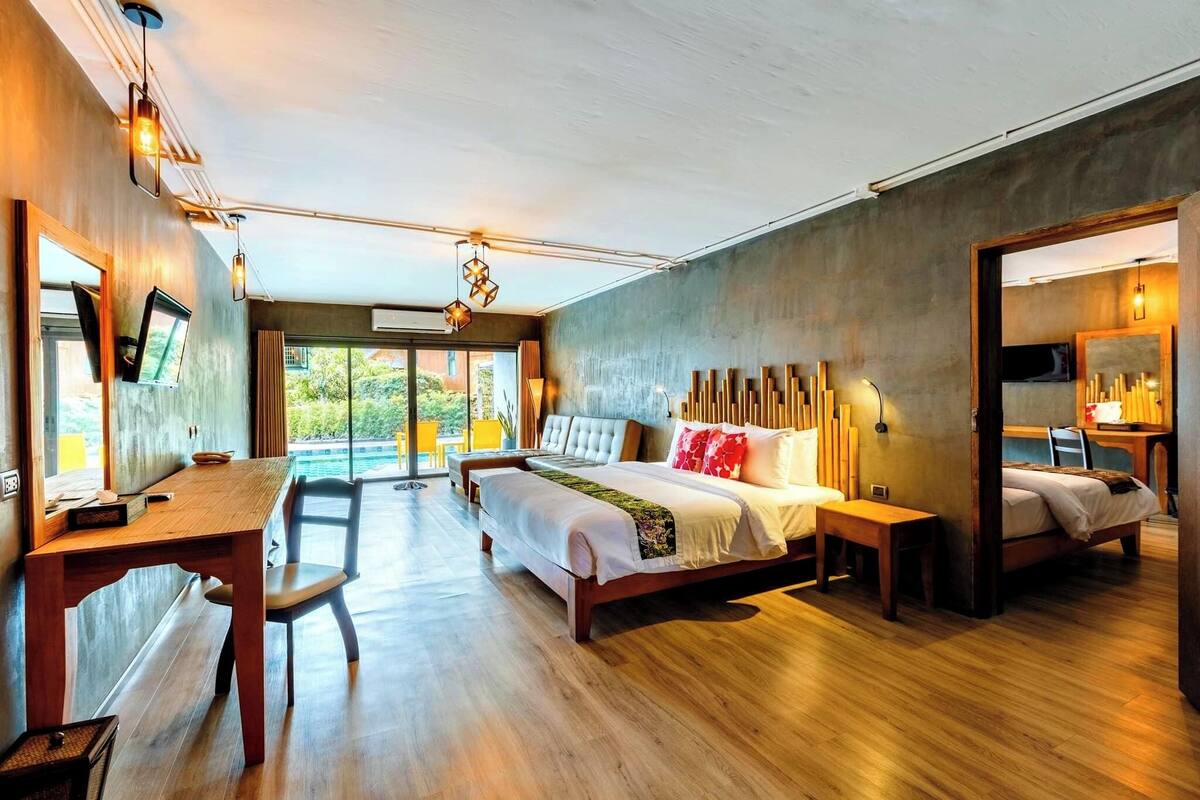 A modern bedroom with a large bed, bedside tables, a desk, and a chair. There is a view of greenery outside the window.