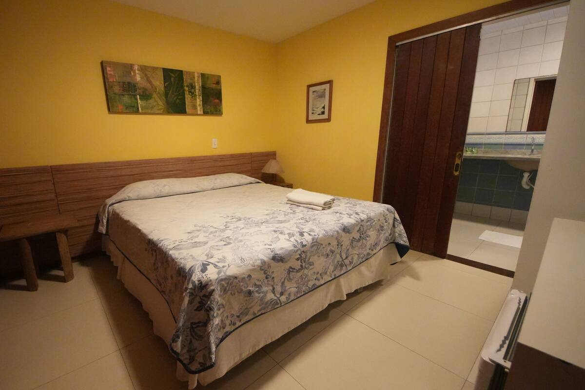 Une chambre à coucher avec un lit, une tête de lit en bois, une table de chevet, une lampe, un tableau et une salle de bain visible à travers une porte.