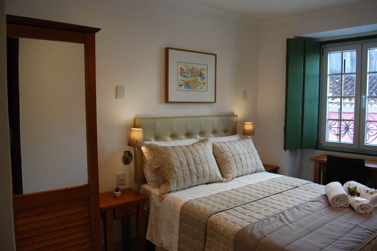 A neatly arranged bedroom with a bed, bedside lamps, a framed picture on the wall, and a window with a view.