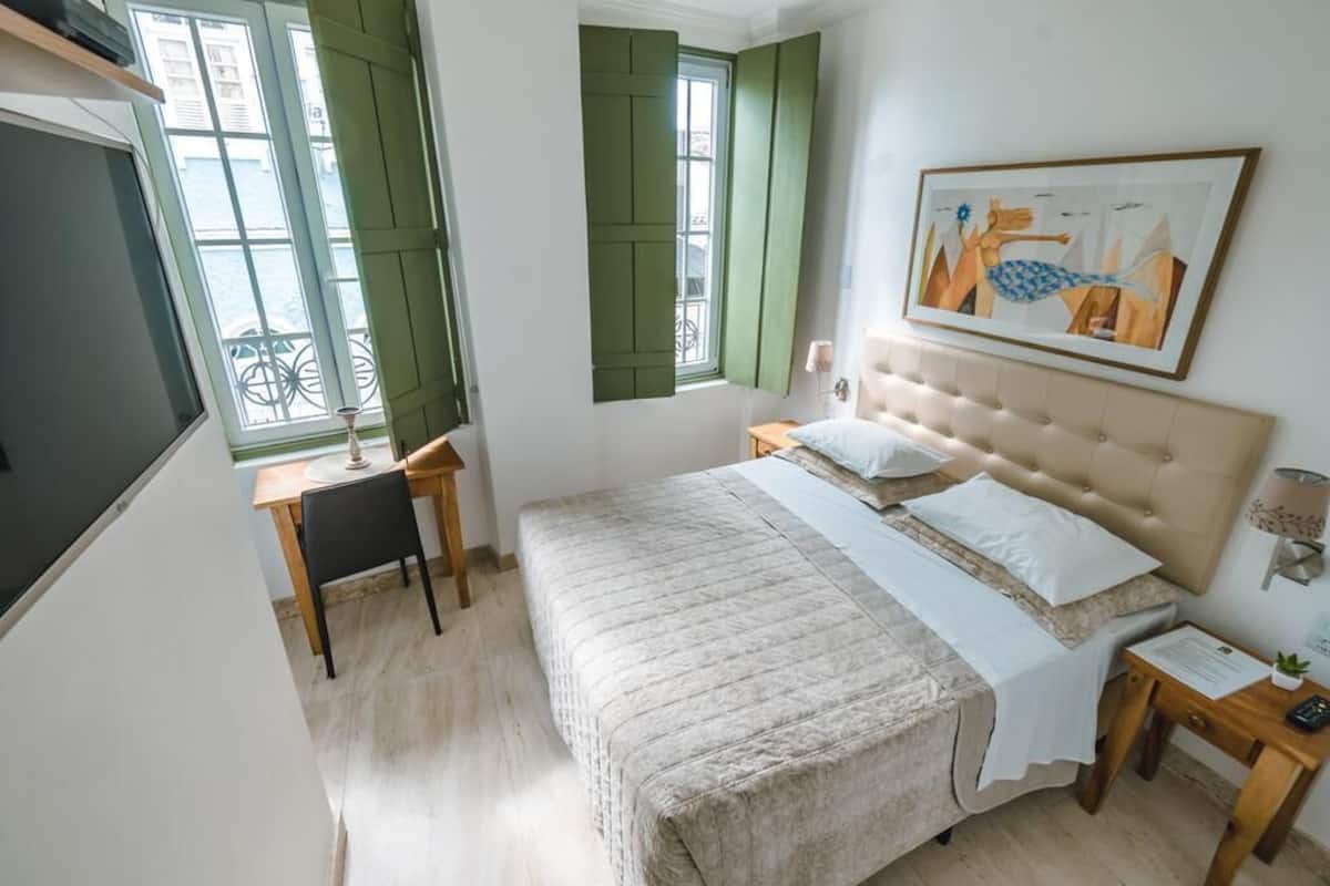 A bedroom with a bed, a nightstand, a chair, a window with shutters, and a painting on the wall.