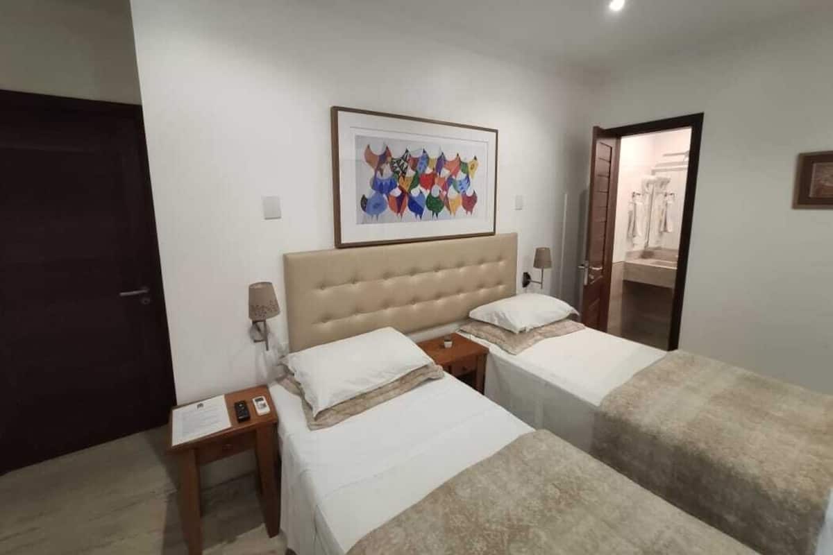 A hotel room with two beds, a large framed artwork above the beds, a door to the bathroom, and a small table with a lamp.