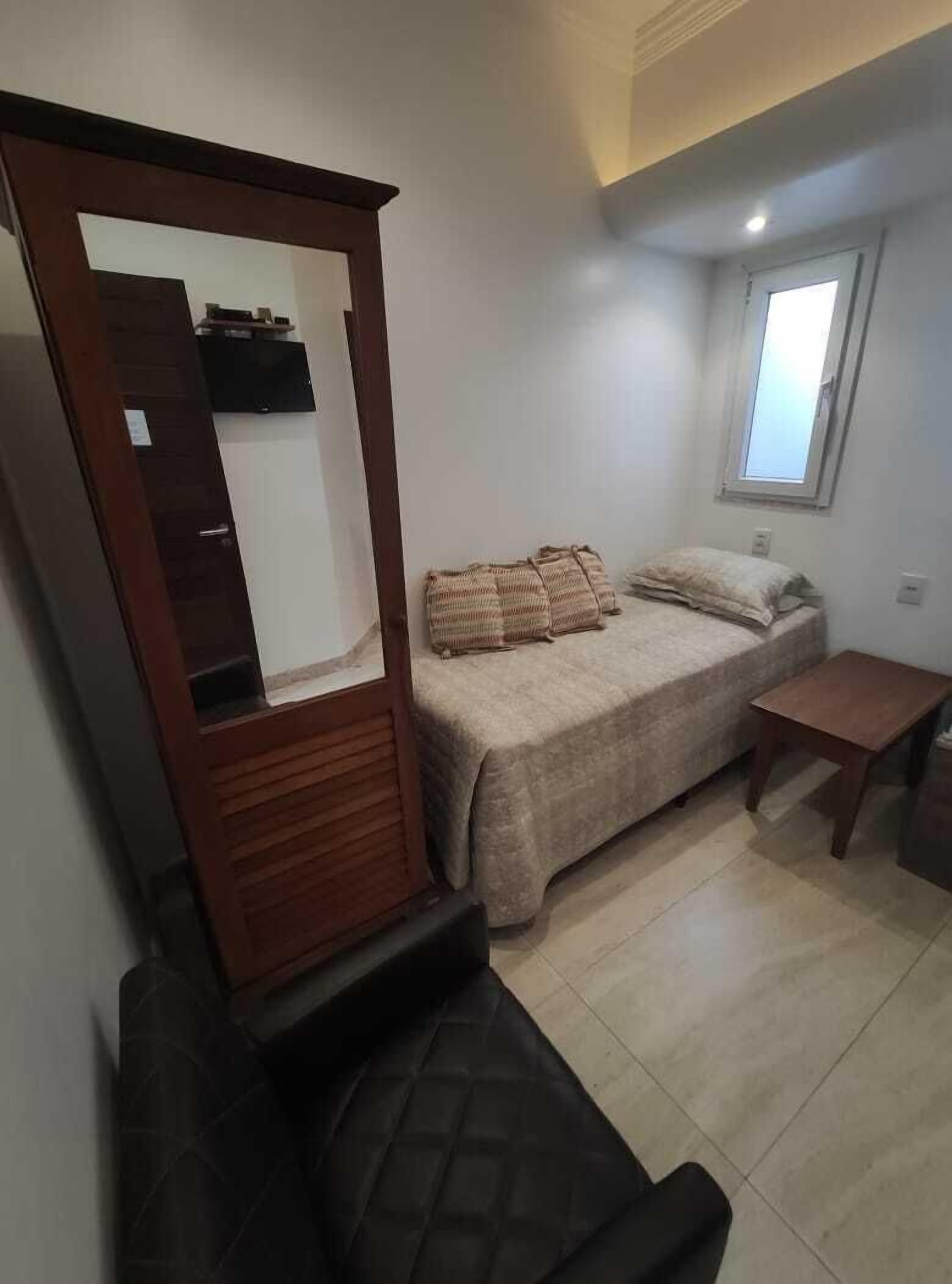 A bedroom with a bed, a wooden wardrobe, a small table, and a window.