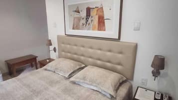 Quarto com uma cama grande, uma obra de arte emoldurada acima da cabeceira, mesa de cabeceira com abajur e uma pequena mesa com telefone e papéis.