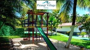 Un parque infantil con un tobogán y estructuras para escalar, rodeado de palmeras y un edificio al fondo.