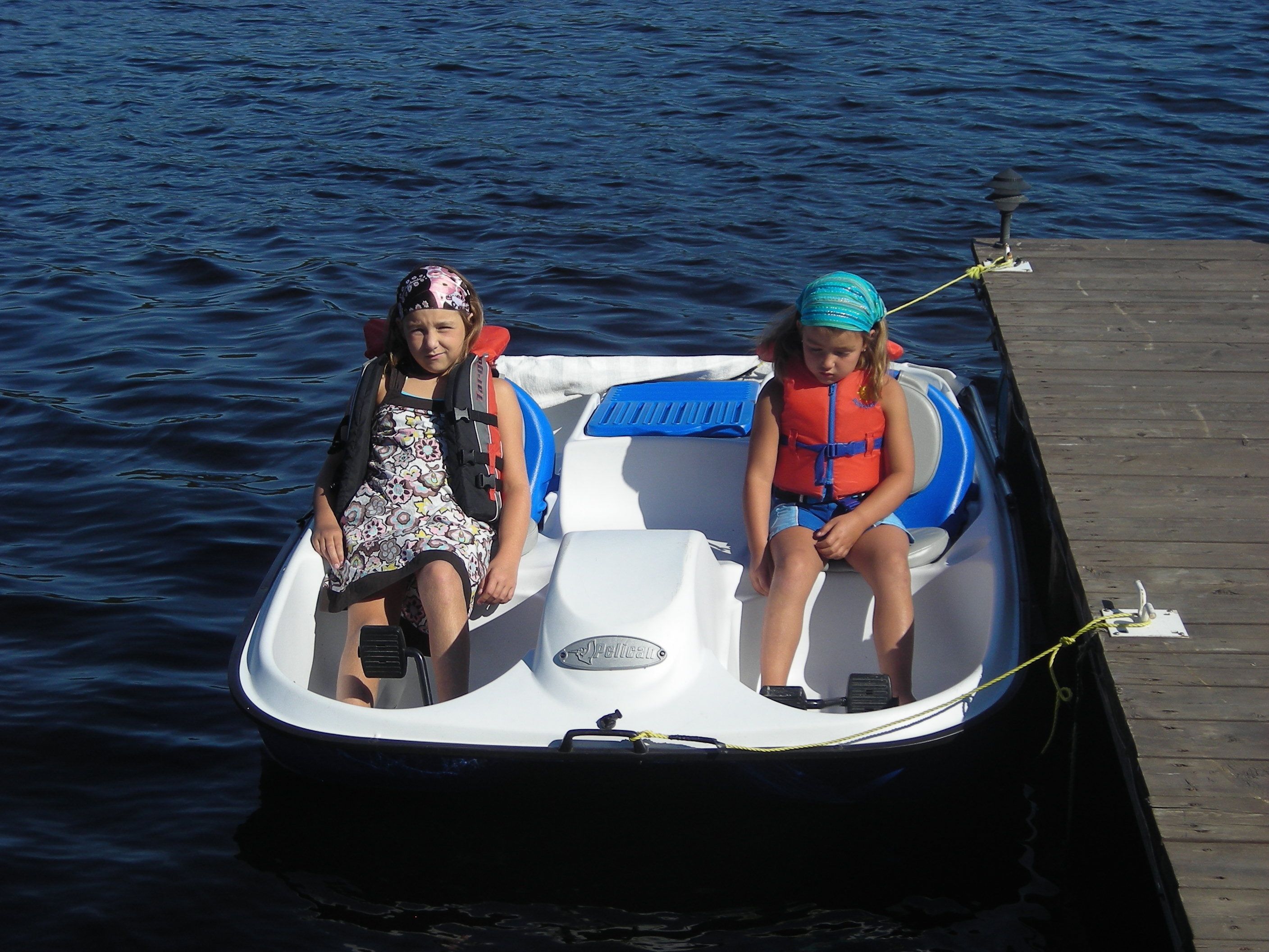 Two children on a small boat with life jackets, docked at a wooden pier.