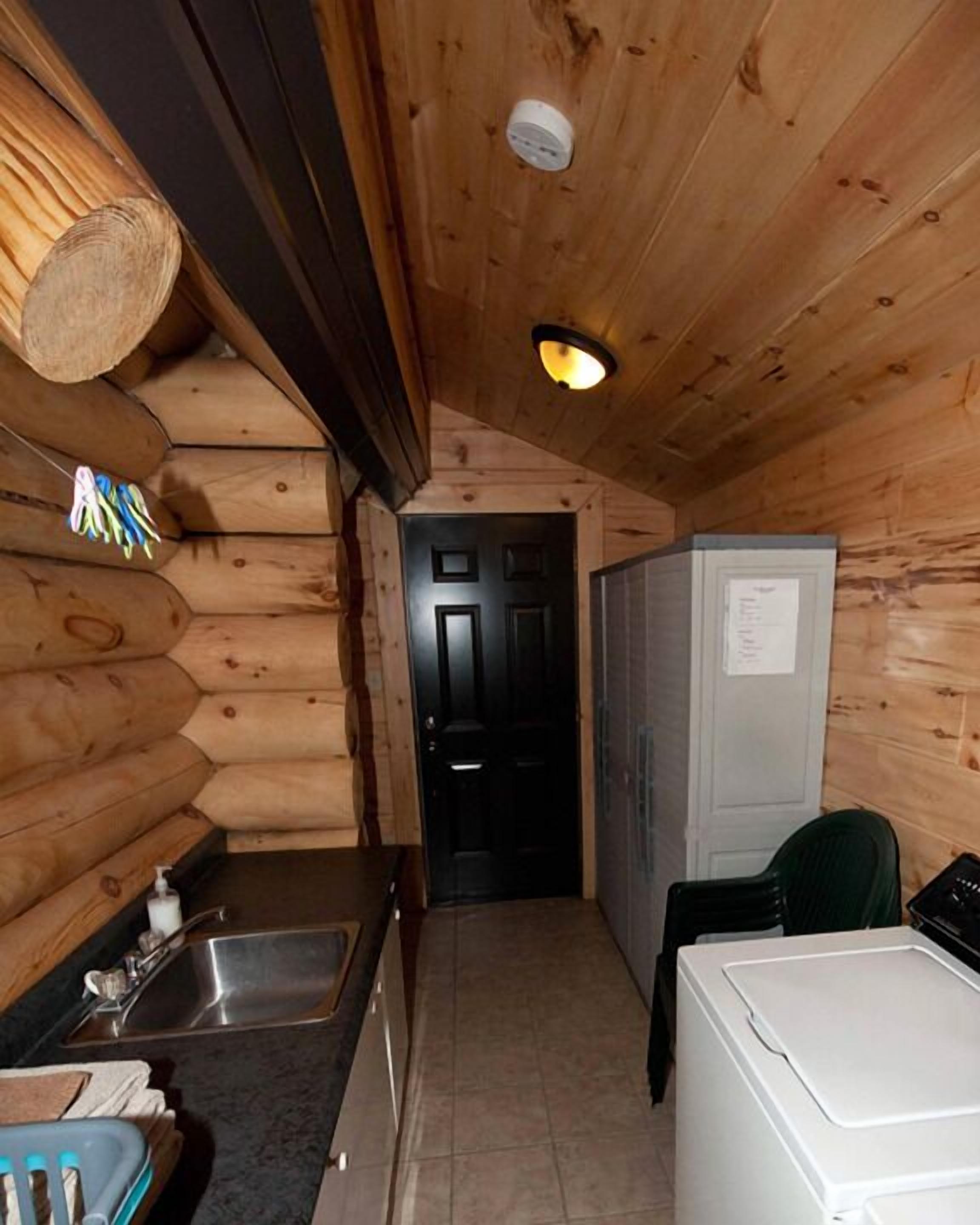 A compact log cabin interior with a kitchen area, including a sink, a refrigerator, and a door leading to another room.