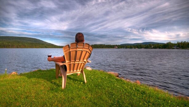A person sitting on a wooden chair by a lake with a view of hills in the background.