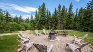 A circular seating area with Adirondack chairs around a fire pit, set in a forested outdoor space with a picnic table. - Le Bois Rond Des Ruisseaux (Saint-Tite-des-Caps)
