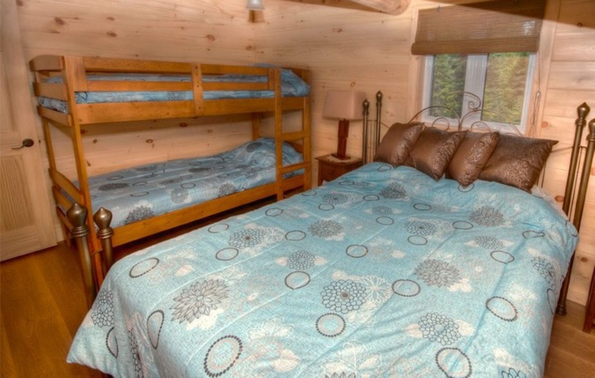 A bunk bed room with a blue patterned bedspread, a wooden bunk bed, a nightstand with a lamp, and a window with a view of trees.