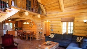 A log cabin interior with a dining area, living room, and a staircase leading to a loft. - Le Bois Rond Des Ruisseaux (Saint-Tite-des-Caps)
