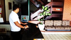 A reception desk with a man at the counter, a large potted plant, a sofa, and a staircase in the background. - Escuna Praia Hotel (João Pessoa)