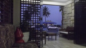 A room with a tiled floor, a wooden bench, a red vase, and a view of parked cars and palm trees outside. - Escuna Praia Hotel (João Pessoa)