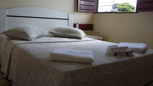 A neatly made bed with patterned bedding, a bedside table with a phone, and a window with shutters. - Escuna Praia Hotel (João Pessoa)