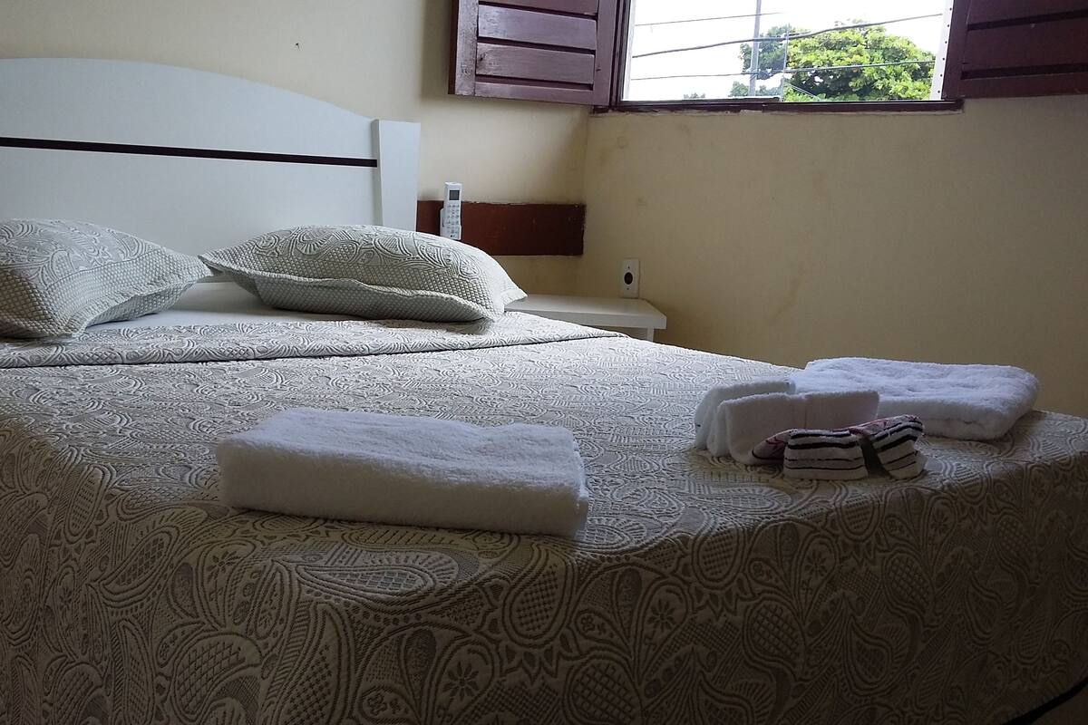A neatly made bed with patterned bedding, a bedside table with a phone, and a window with shutters.
