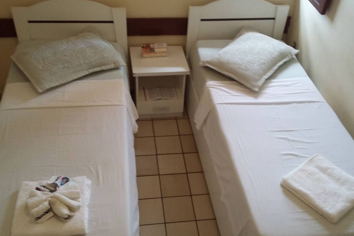 Two single beds with white linens and pillows, a small bedside table with a book, and a tiled floor.