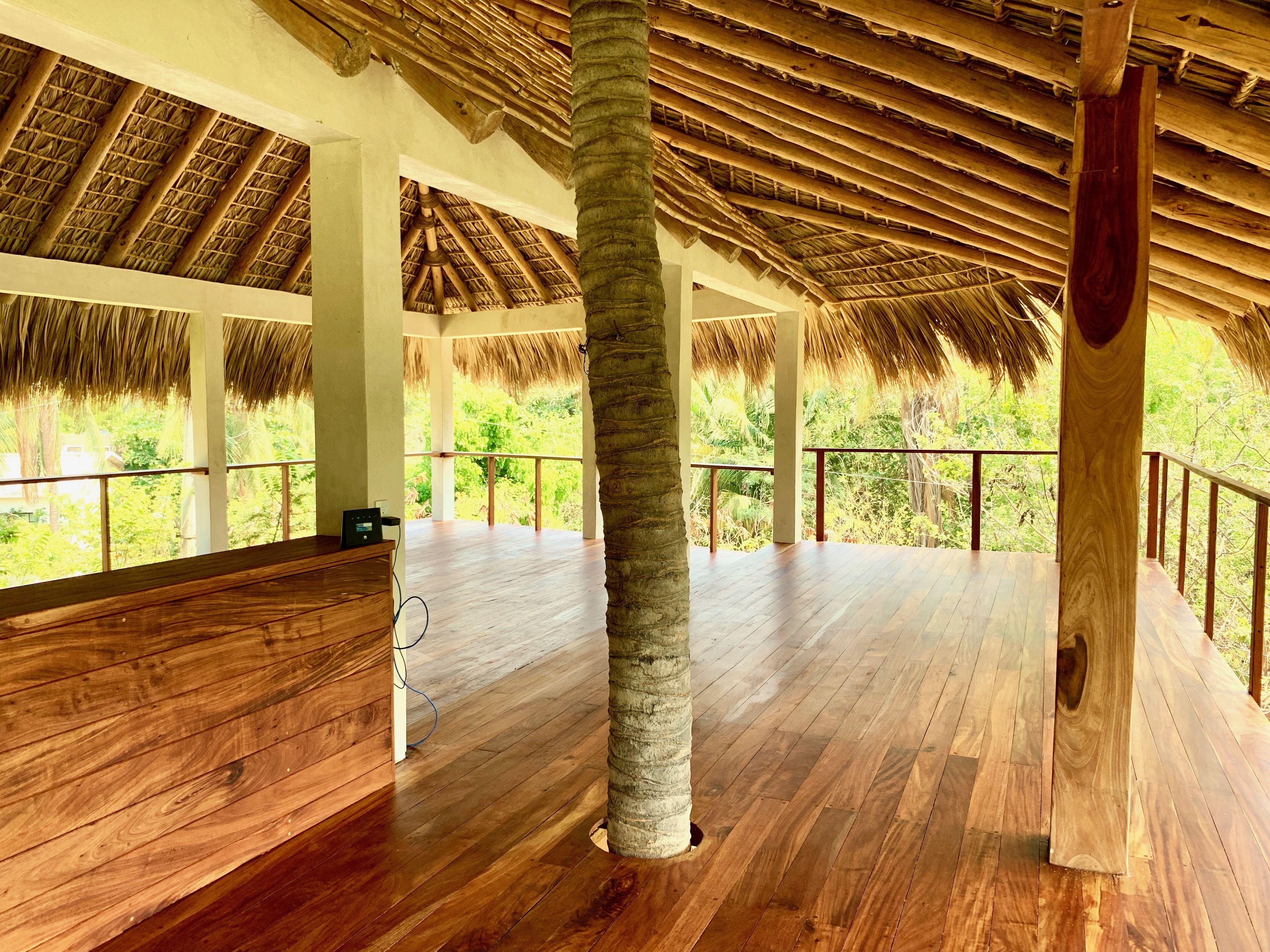 A spacious room with wooden flooring, a thatched roof, and a view of lush greenery outside.