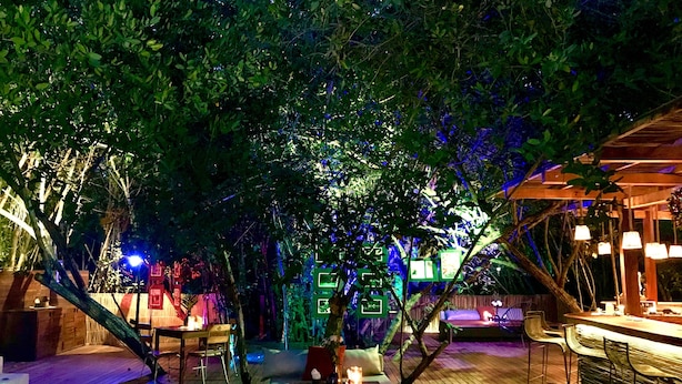 An outdoor seating area with tables and chairs, surrounded by trees and illuminated by ambient lighting.