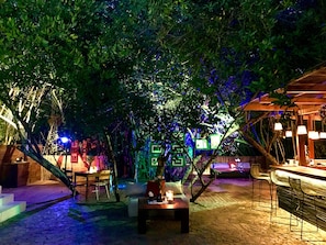 Un espacio al aire libre con mesas y sillas, rodeado de árboles e iluminado con luz ambiental.