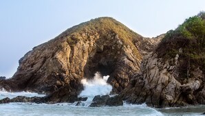 A rocky coastal landscape with a cave entrance, waves crashing against the shore, and a sandy beach.