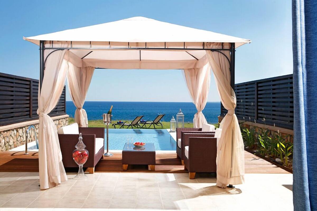 Un área de estar al aire libre con techo, piscina, sillones de descanso y vistas al océano.