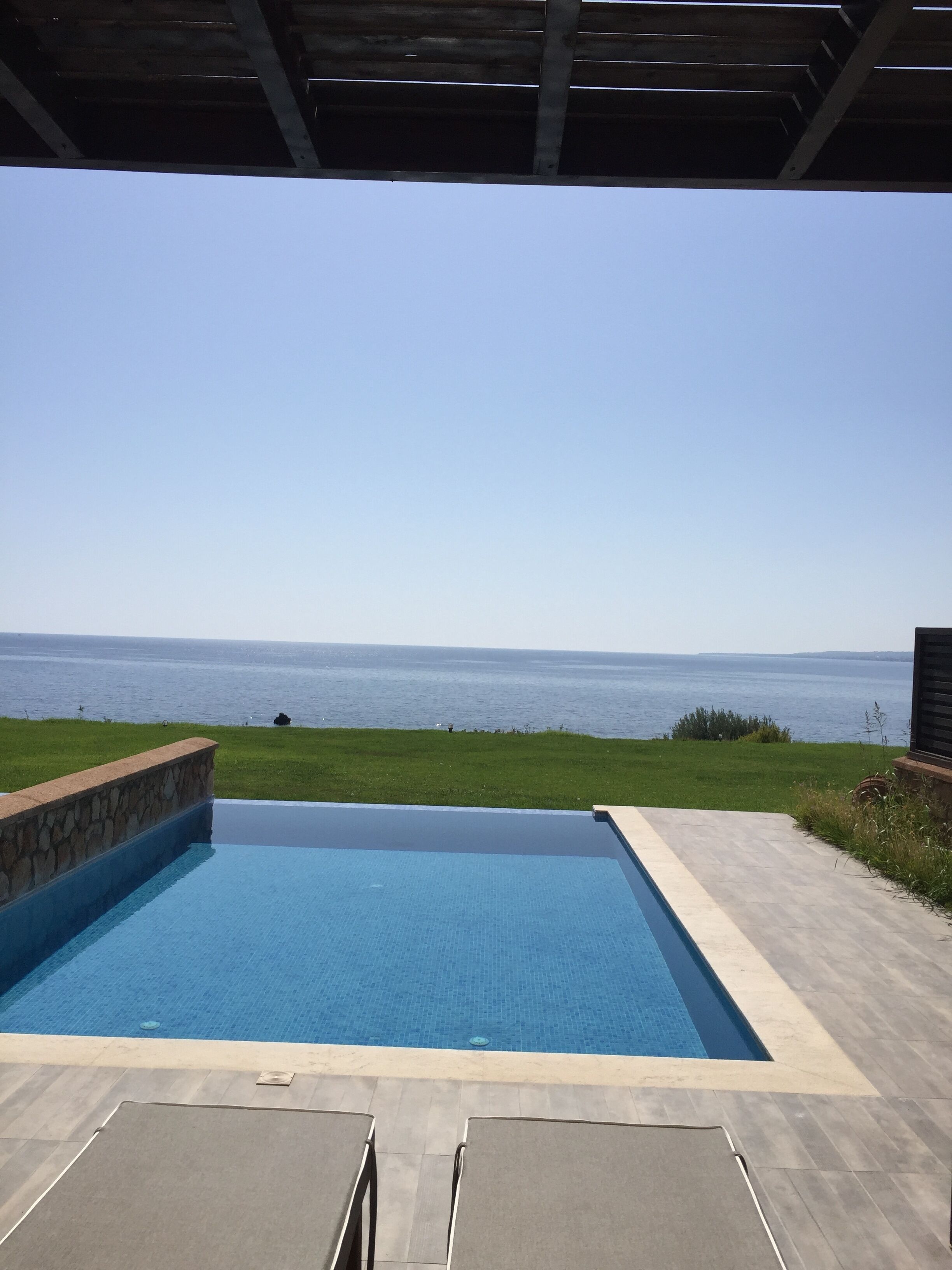 A swimming pool with a view of the sea, surrounded by a stone wall and a wooden pergola.