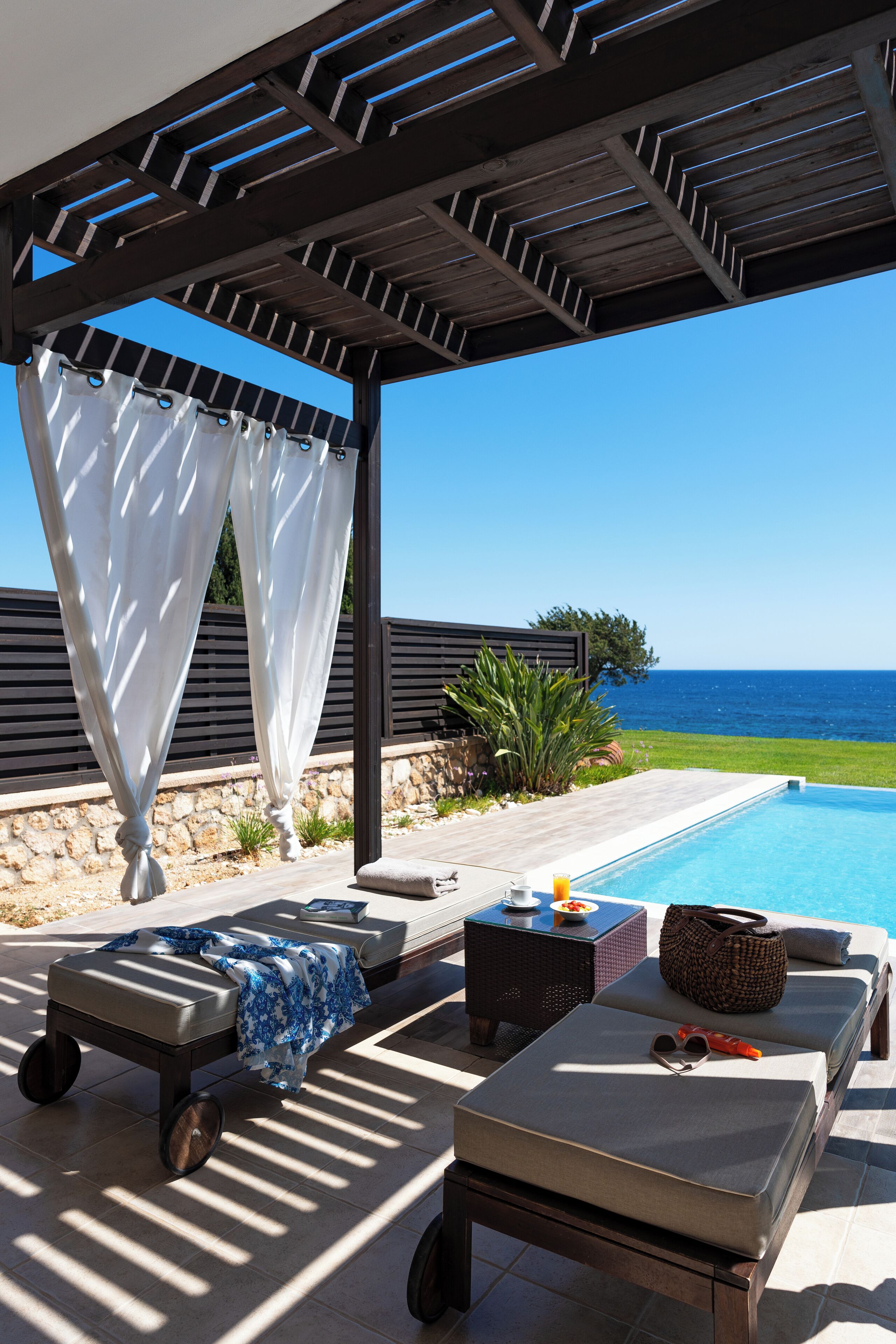 Un área de descanso junto a la piscina con vistas al océano, una zona de estar cubierta y una pequeña mesa con una bebida.