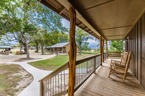 Een houten veranda met een balustrade, een schommelstoel en uitzicht op een camping met hutten en bomen.