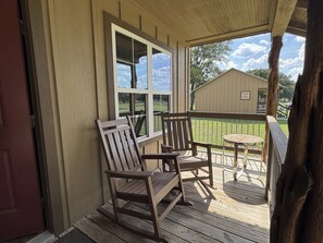 Une véranda en bois avec deux fauteuils à bascule et une petite table, donnant sur une cour verdoyante et un bâtiment.