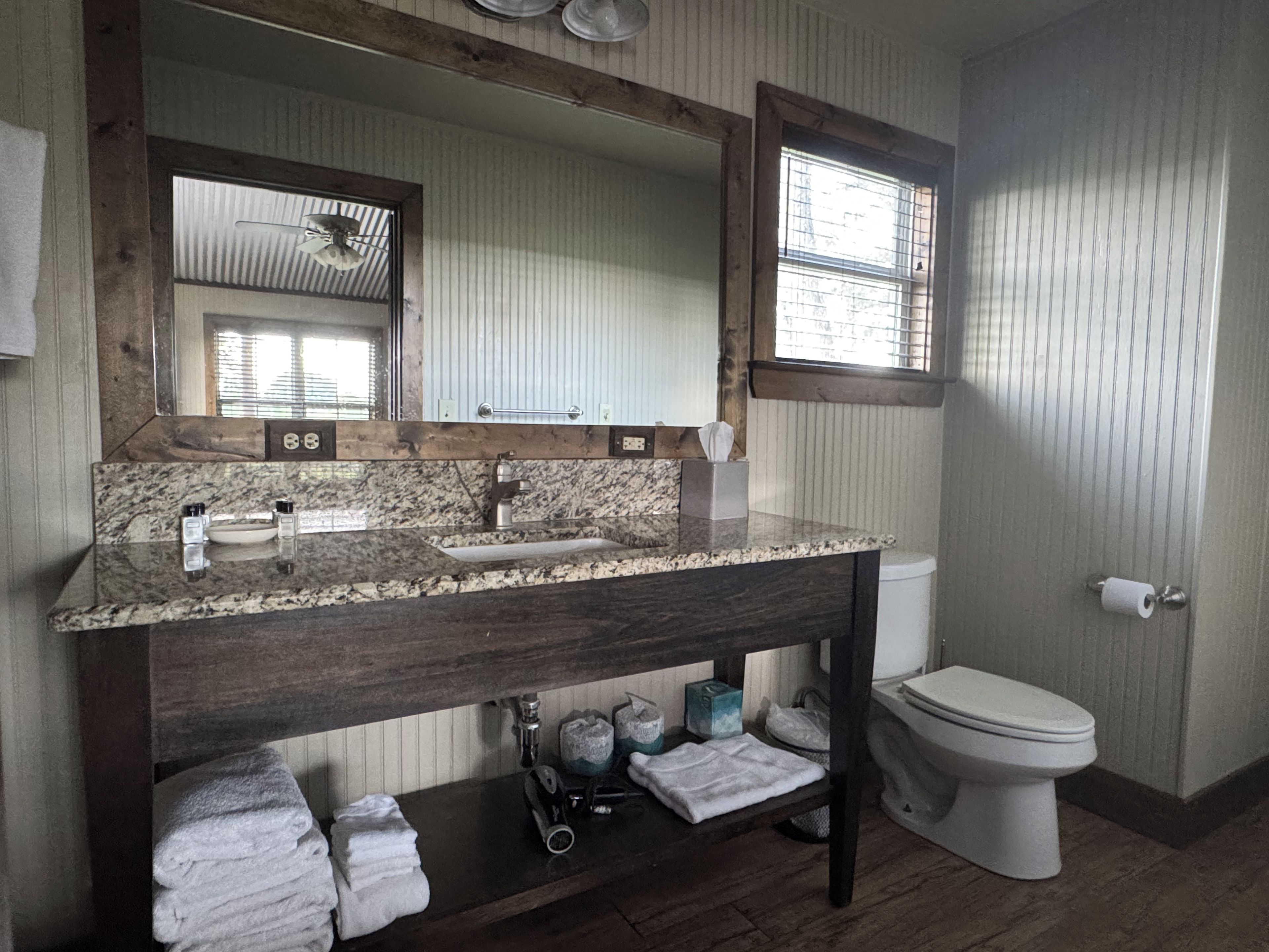 Une salle de bain avec un meuble-lavabo en bois, un évier, un miroir, des toilettes et une fenêtre avec des stores.