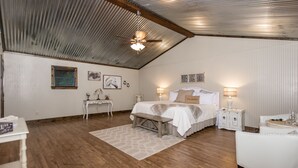 Un dormitorio con techo inclinado, una cama con cabecera, mesitas de noche, un banco y una pequeña mesa con una lámpara.