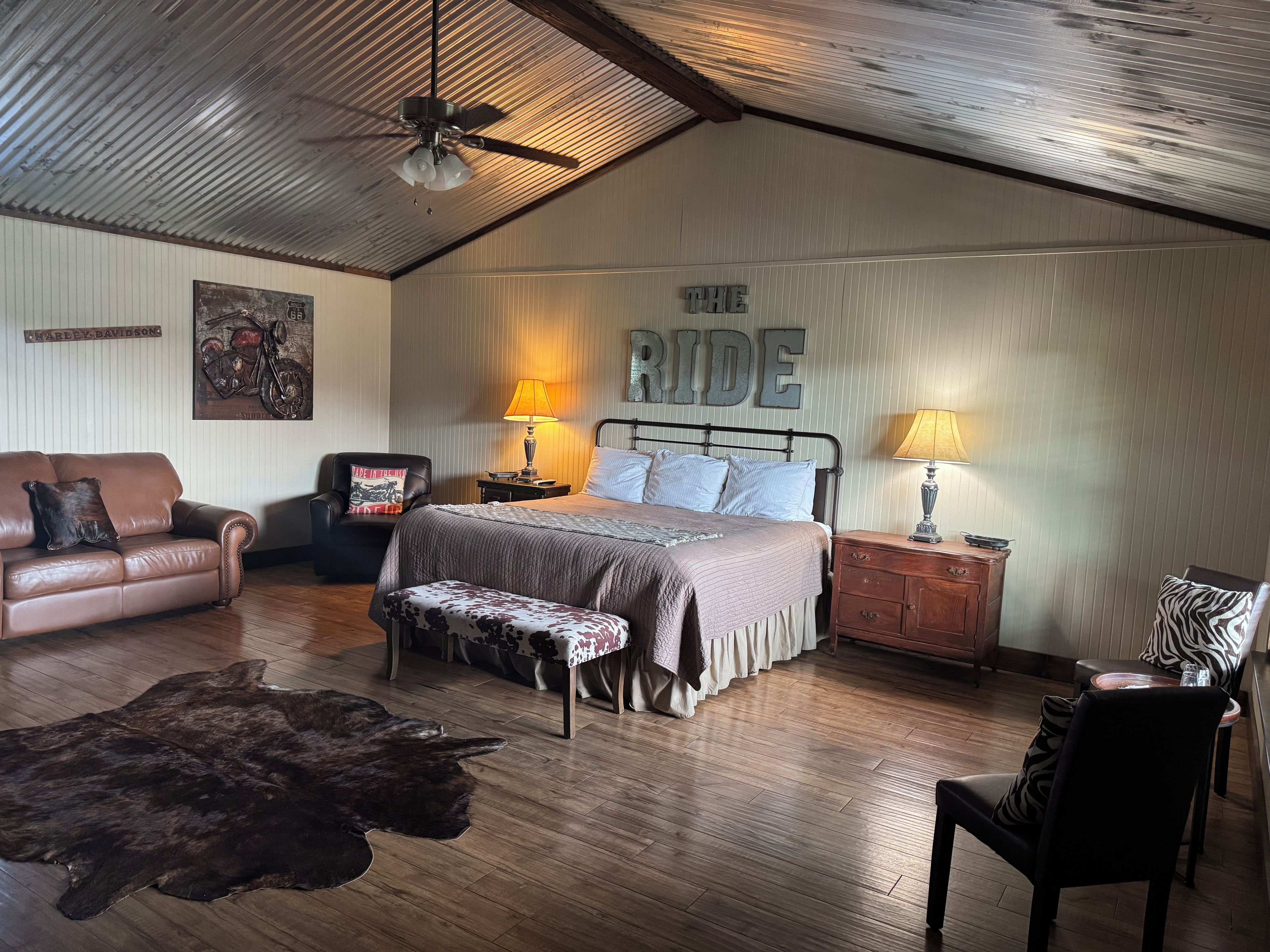 A room with a bed, a leather sofa, a wooden floor, a ceiling fan, and a wall with 'THE RIDE' sign.