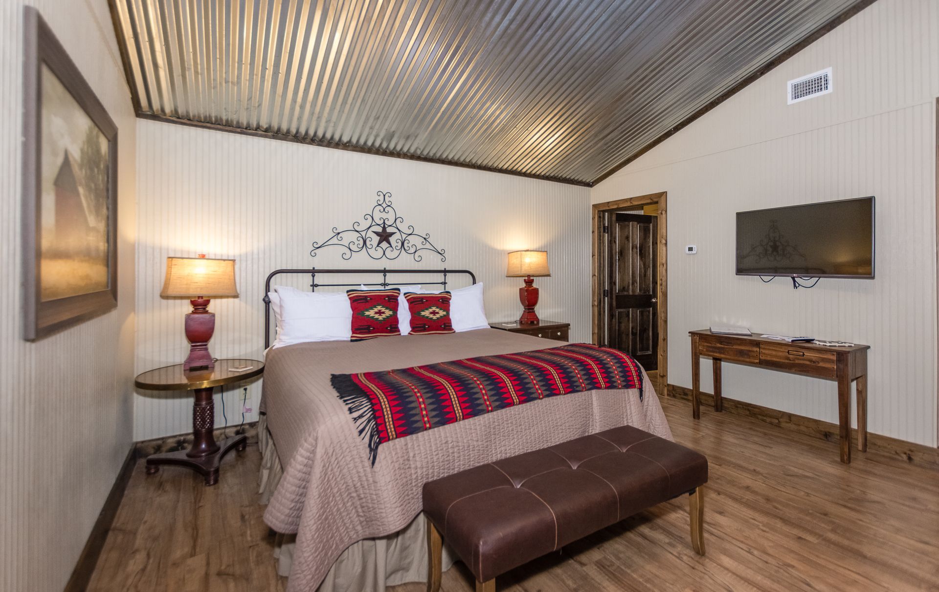 A bedroom with a bed, bedside lamps, a television, a wooden bench, and a door.
