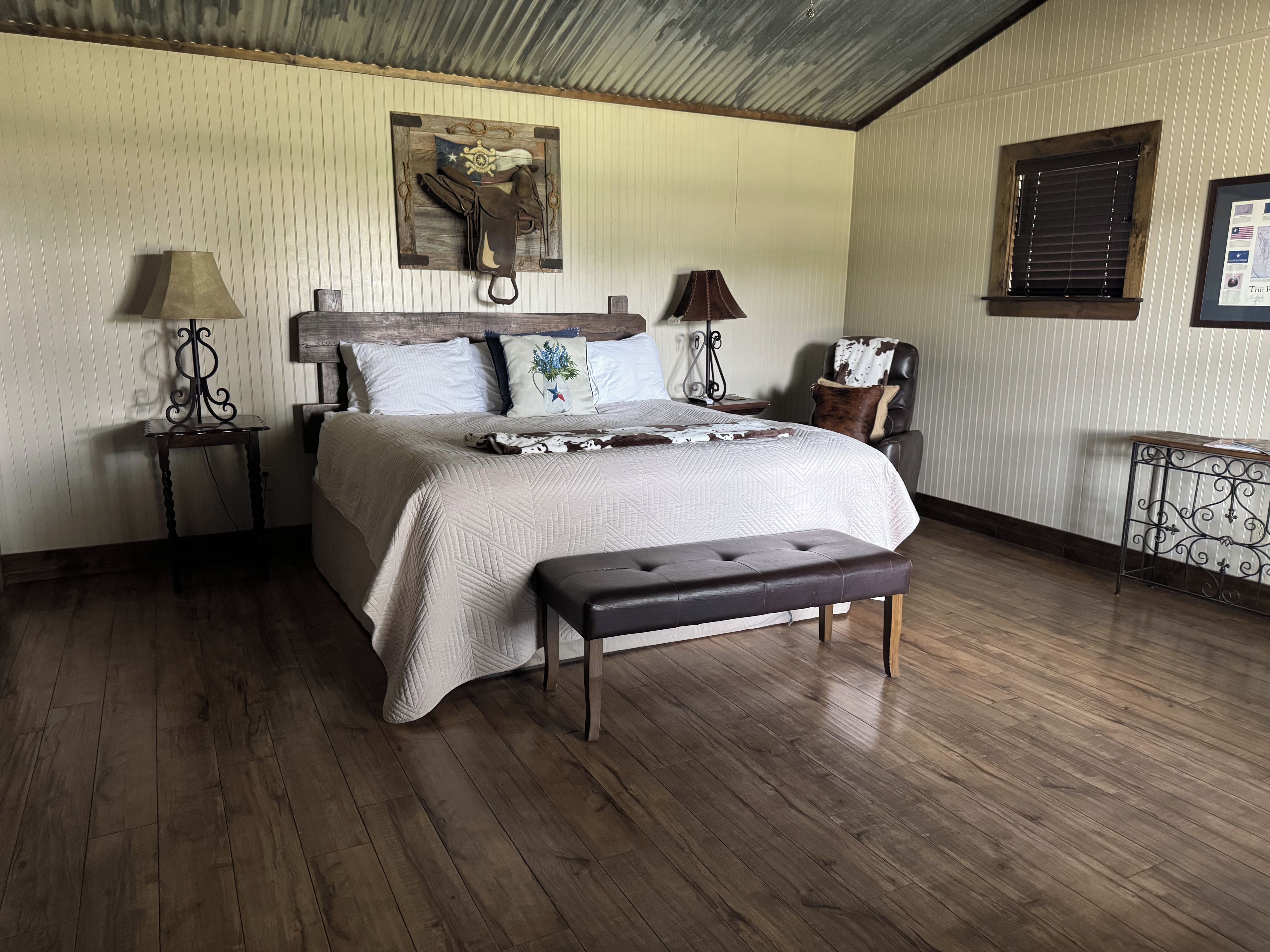 Ein Schlafzimmer mit einem großen Bett, zwei Nachttischen, einem Sessel, einer Bank und einem an der Wand hängenden Kunstwerk.