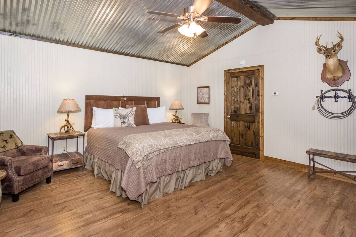 Un dormitorio con una cama de madera, dos lámparas de noche, una silla de madera, un banco de madera, un ventilador de techo y una cabeza de venado montada en la pared.
