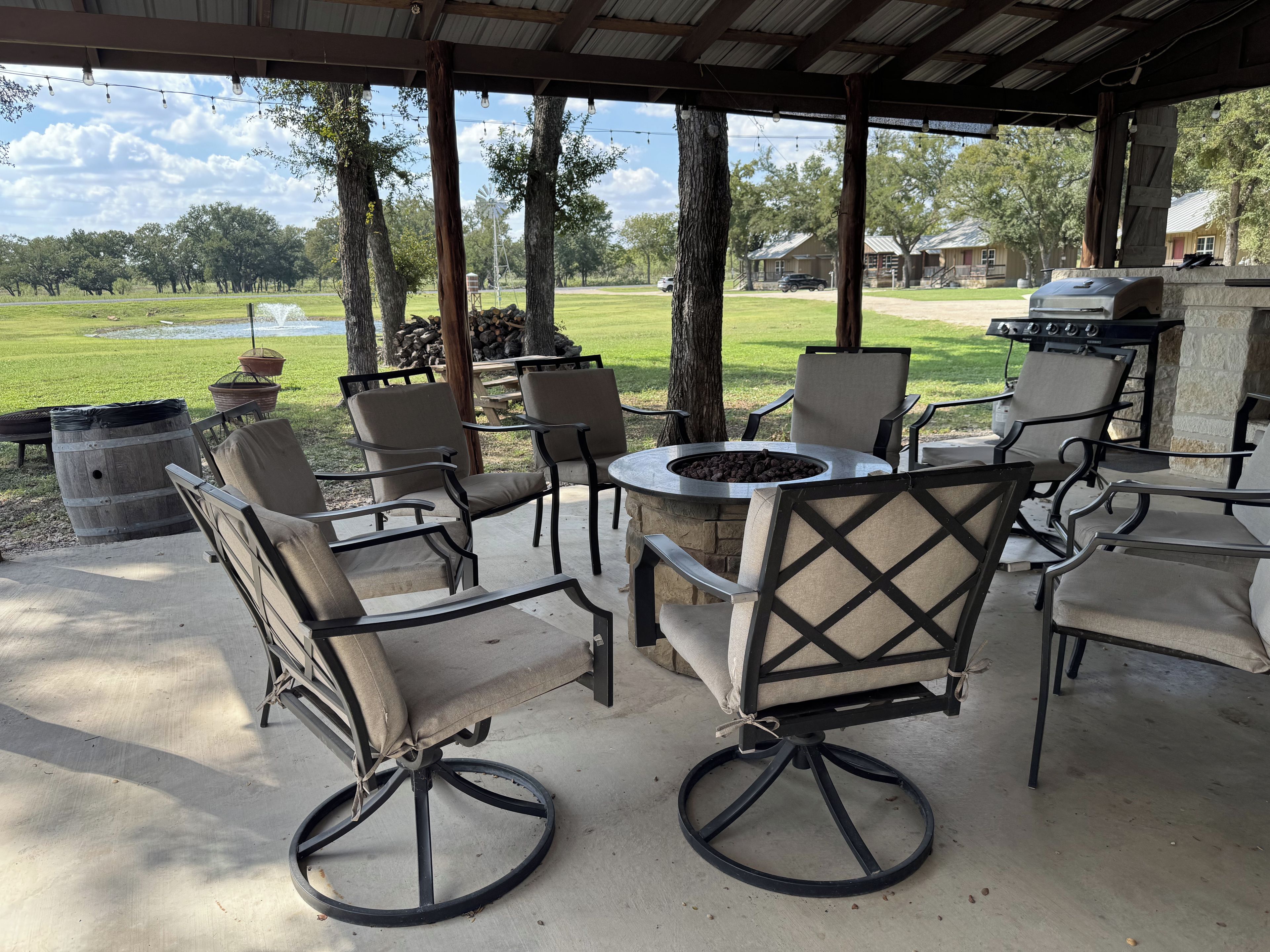 An outdoor patio with a fire pit table surrounded by chairs, a grill, and a view of a grassy field.