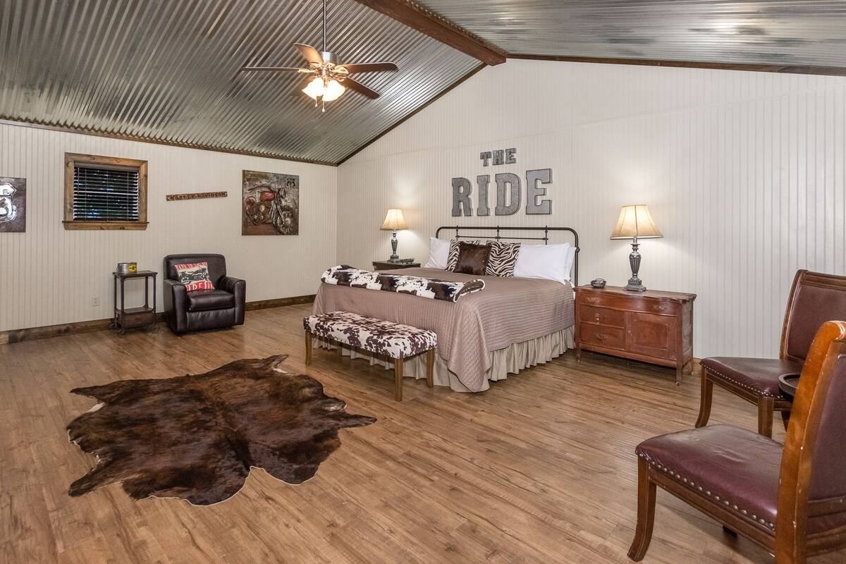 Un dormitorio de estilo rústico con cama, un sillón de cuero, un banco de madera, una mesita y un ventilador de techo fijado a la pared.