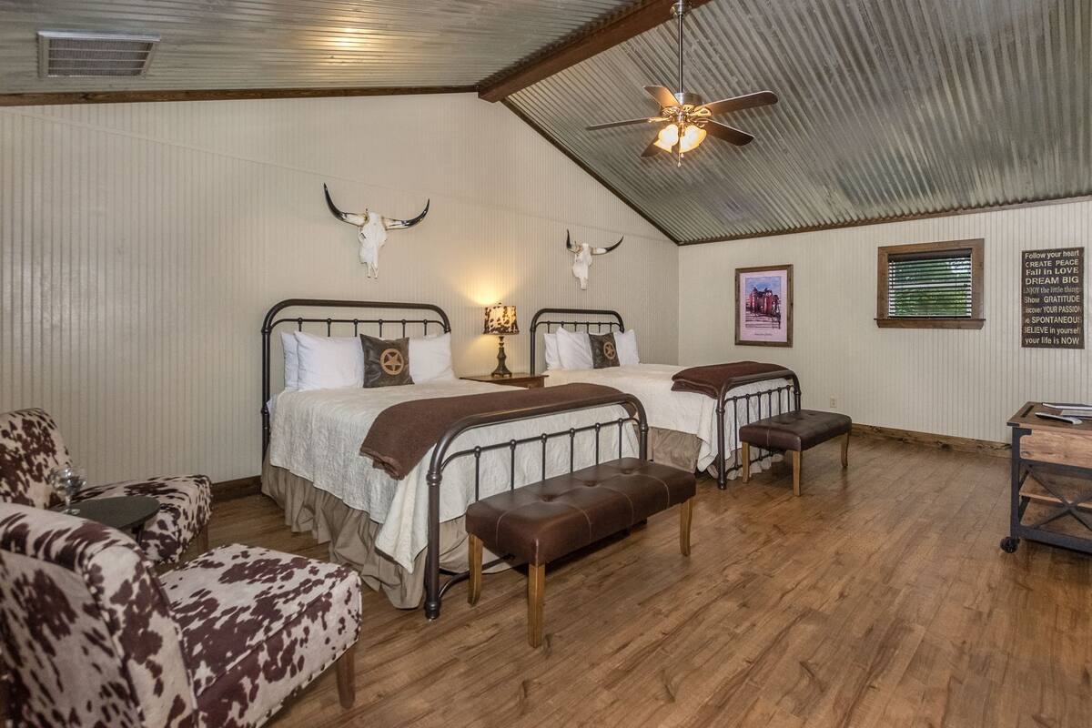 Una habitación rústica con dos camas, un ventilador de techo, una decoración con cráneo de vaca, un cuadro enmarcado y una cita motivacional en la pared.
