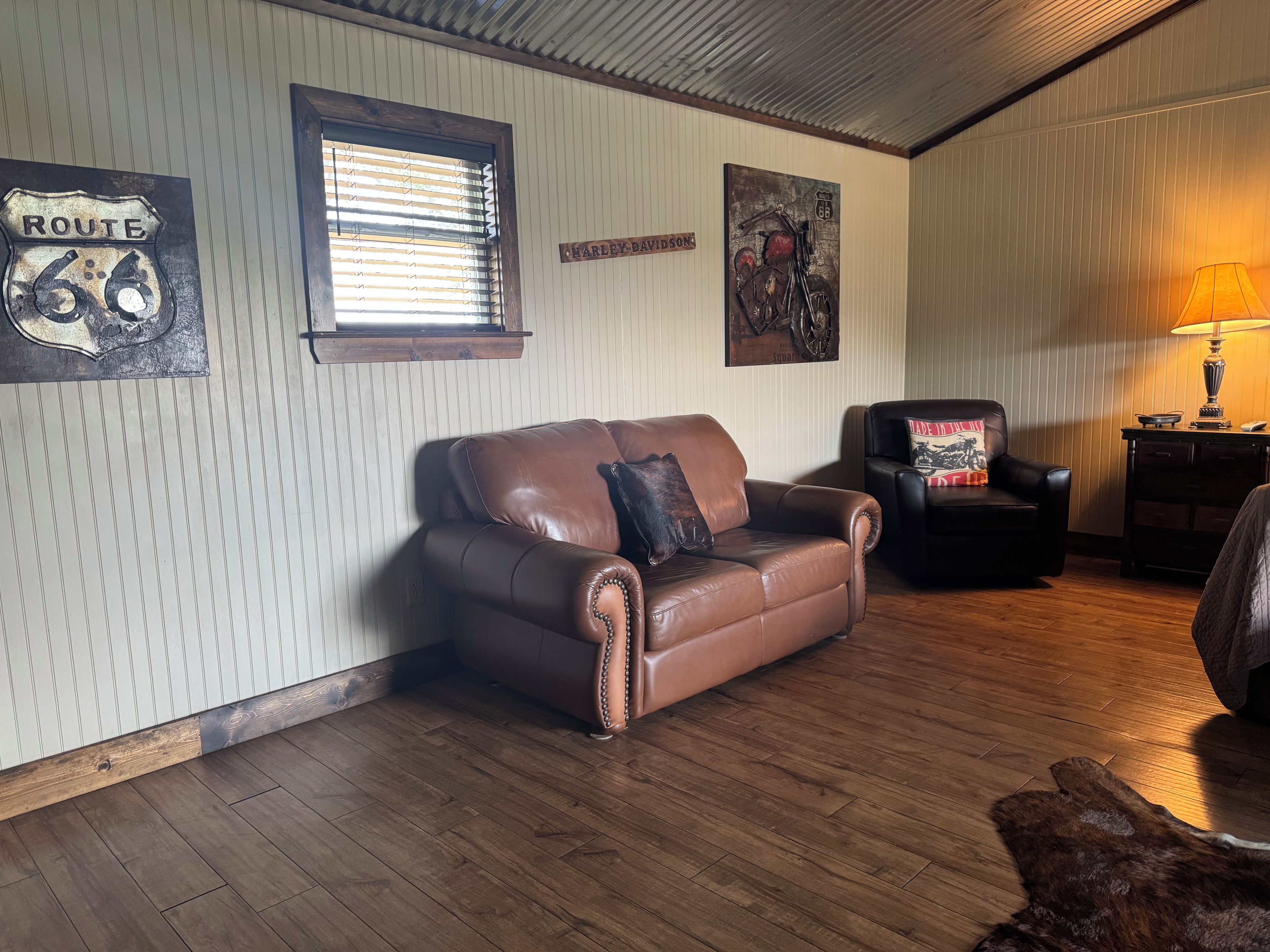 A room with a brown leather sofa, a black leather armchair, a wooden floor, a window with blinds, and wall decorations including a Route 66 sign and a Harley-Davidson sign.
