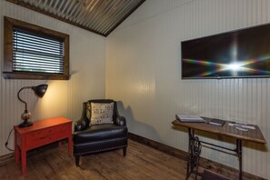 A room with a wooden floor, a black leather chair with a cushion, a desk with papers, a lamp, and a window with blinds. - Vineyard Trail Cottages - Adults Only (Fredericksburg)