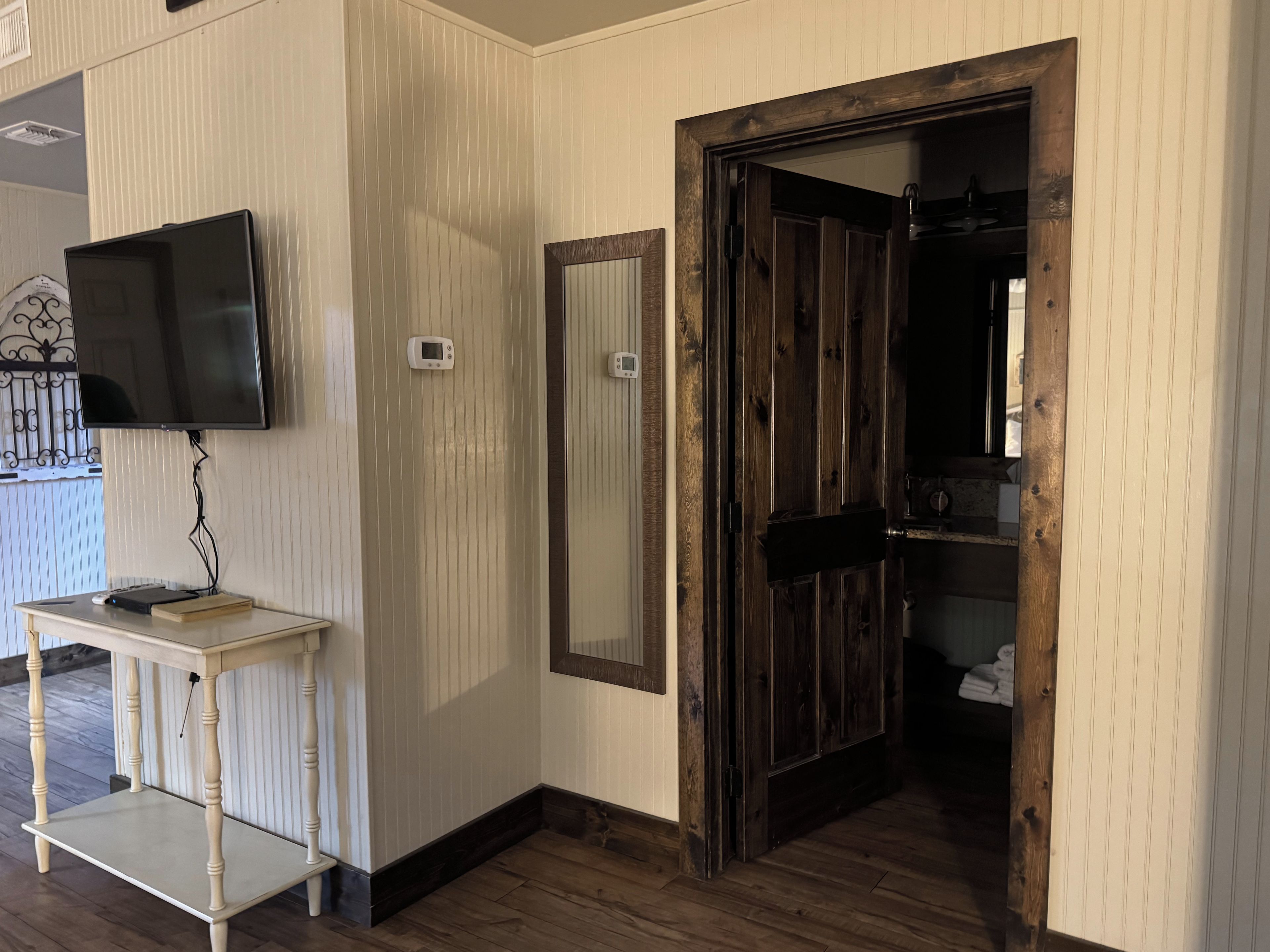 A room with a television mounted on the wall, a small table with a book, a mirror, and a wooden door leading to another room.