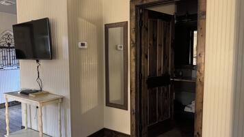 A room with a television mounted on the wall, a small table with a book, a mirror, and a wooden door leading to another room.