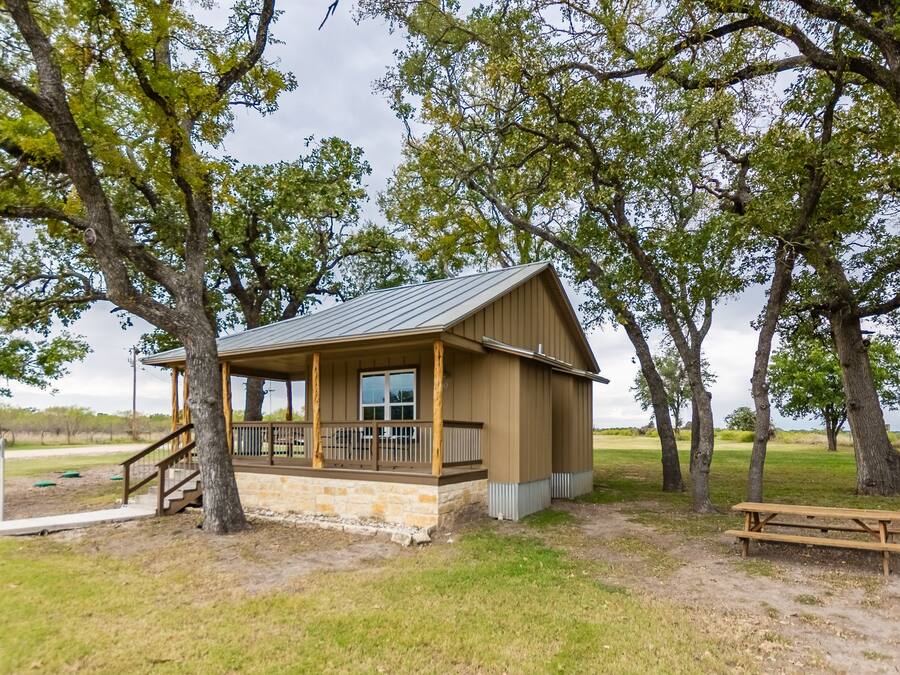 A small wooden cabin with a porch, surrounded by trees in a grassy area.