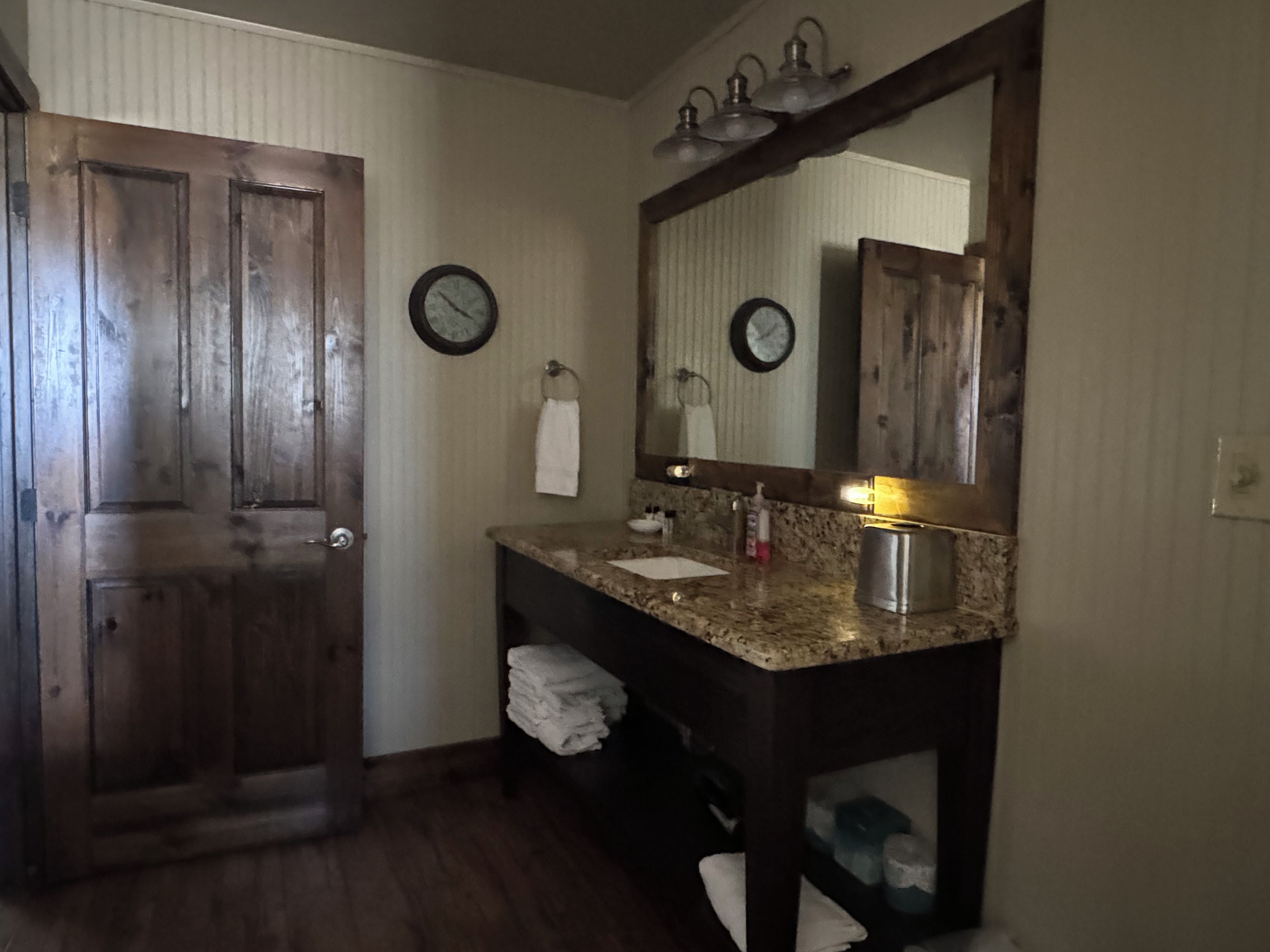 A bathroom with a wooden door, a sink with a mirror, and two wall clocks.