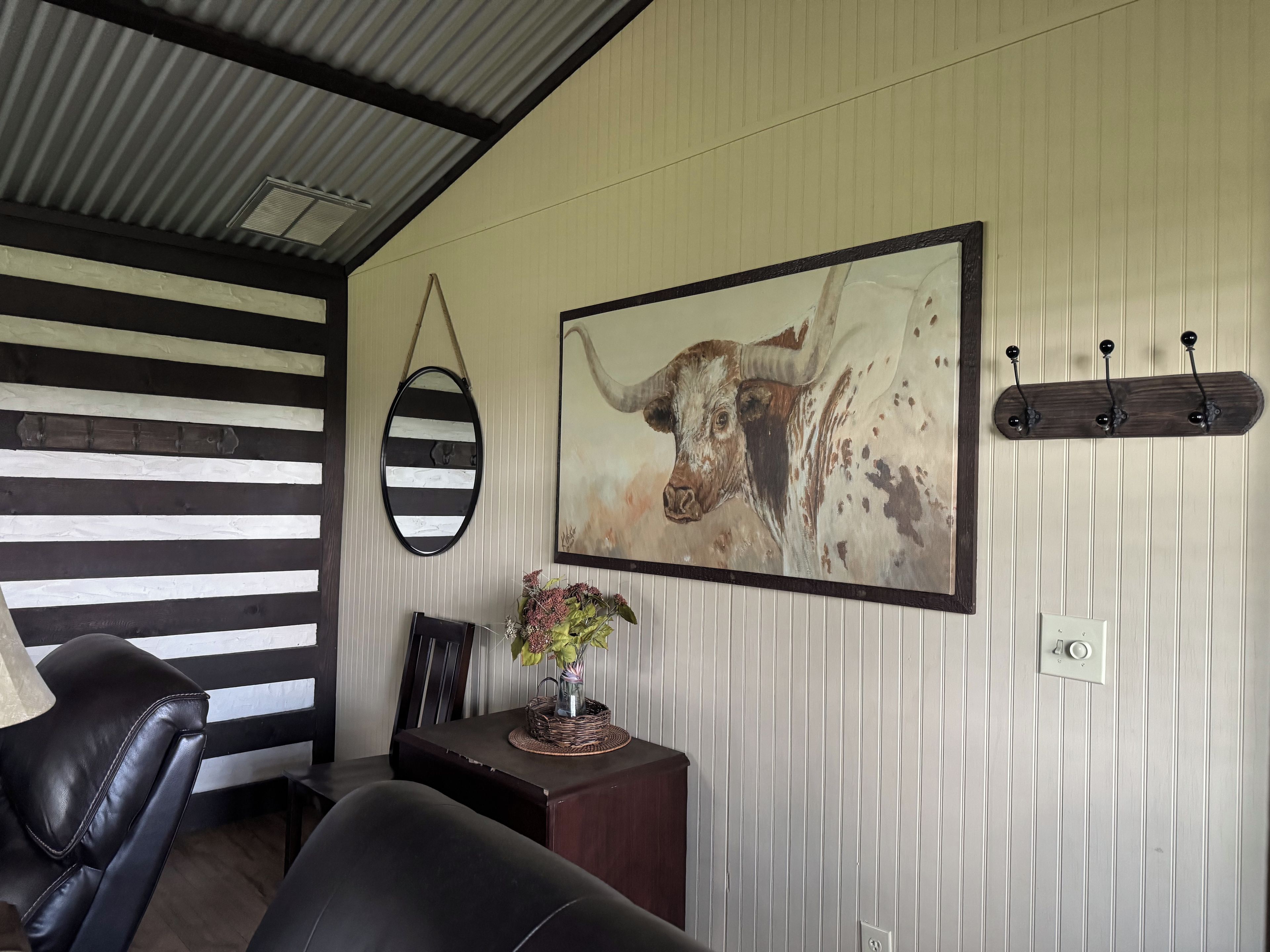 Une pièce avec un mur à rayures, un tableau encadré représentant une vache, un porte-manteau, une lampe et une petite table sur laquelle est posé un vase de fleurs.