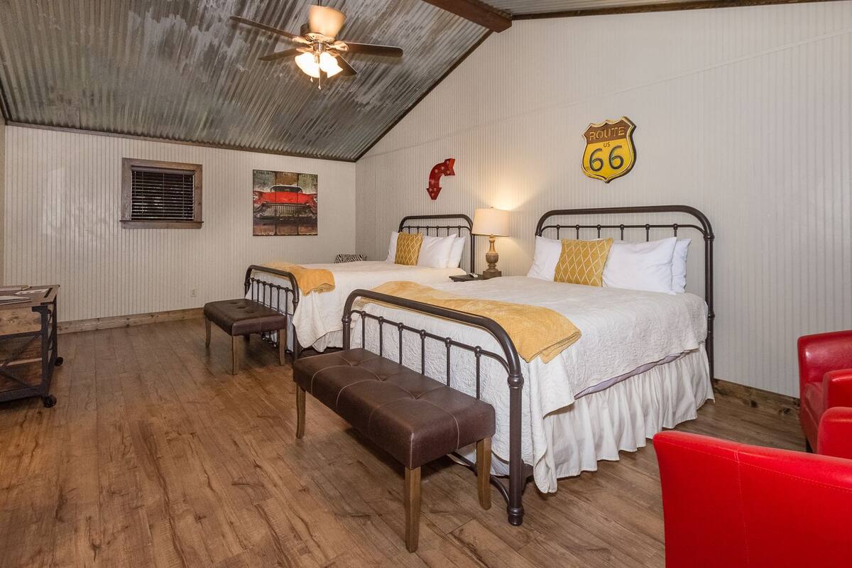 Una habitación con dos camas, un banco, una silla roja, una lámpara y un cartel de Route 66.
