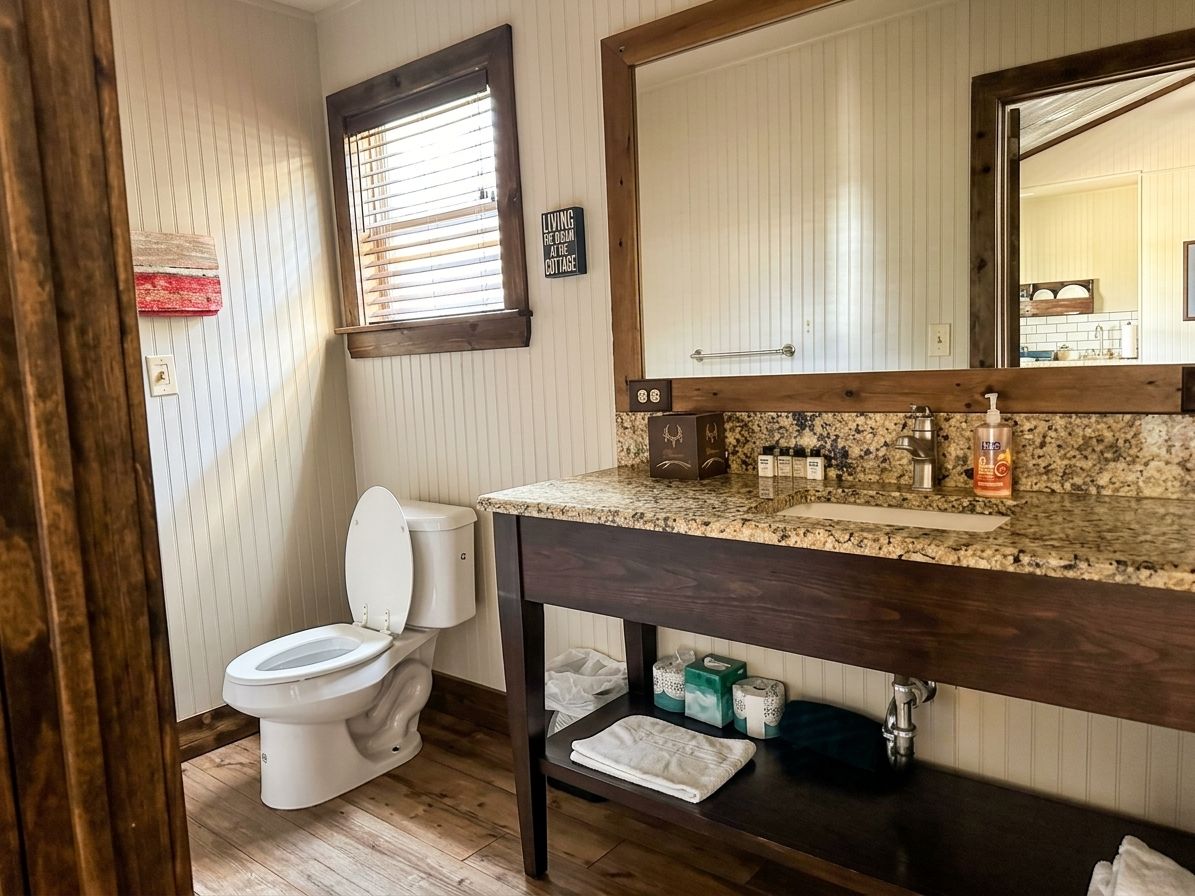 Une salle de bain comprenant des toilettes, un meuble-lavabo avec un miroir et une fenêtre avec des stores.
