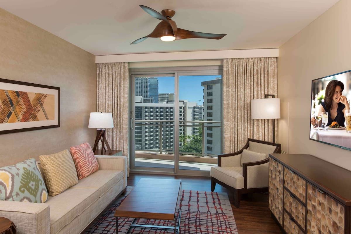 Ein Hotelzimmer mit Sofa, Sessel, Fernseher, Deckenventilator und einem großen Fenster mit Blick auf die Stadt.
